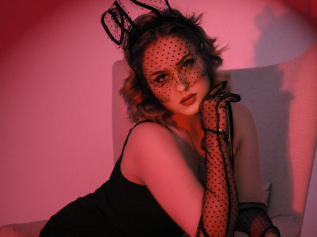 Lisa Francis, avec un masque à pois, une pièce de tête en dentelle noire et des gants assortis, portant une robe noire, a sa main sur son menton, sous un éclairage rouge, et regarde séductrice l'appareil photo..