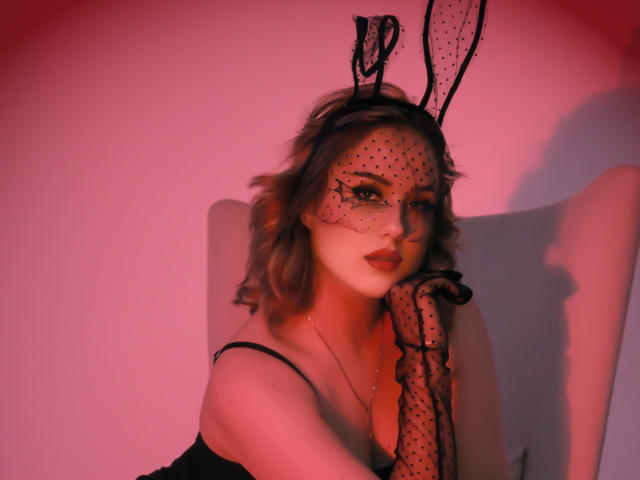LisaFrancis with wavy brown hair, wearing black polka-dot mask and bunny ears, red lipstick, black strap dress, and black polka-dot glove, in pink-red lighting.