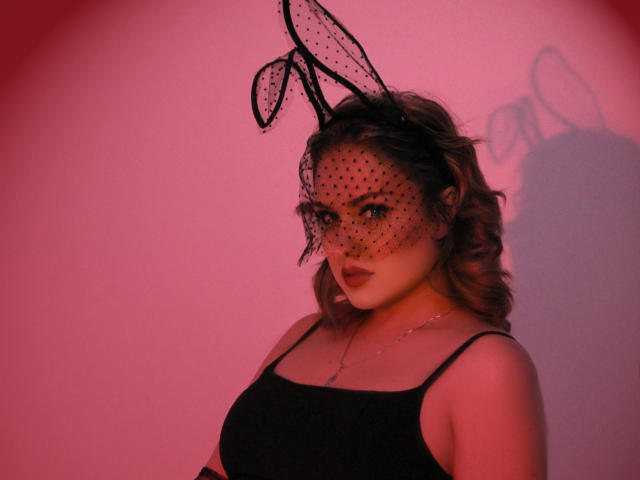 LisaFrancis with wavy brown hair, black sheer mask with polka dots and bunny ears, wearing a black spaghetti strap top, red-lit shadow on wall, looking seductive.