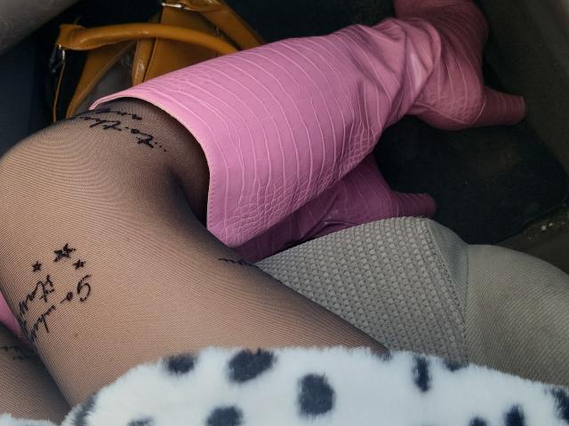 La jambe de Choukette, couverte de bottes roses, est pliée, révélant un texte noir tatoué sur des collants transparents, avec un sac jaune et une couverture blanche à pois en arrière-plan..