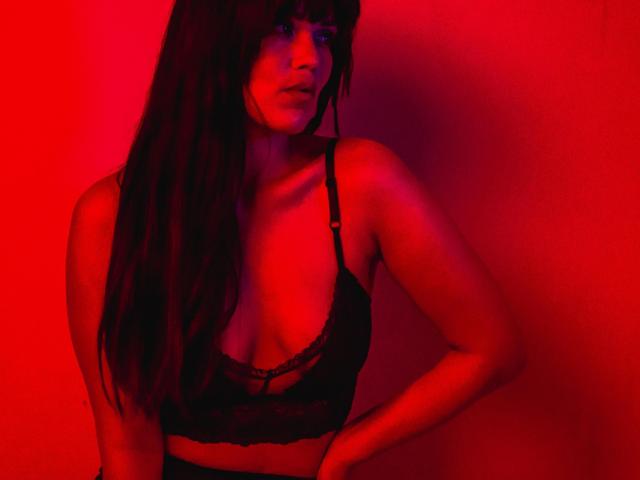 MissNight, con lunghi capelli scuri, indossa un reggiseno di pizzo nero, illuminata di rosso da sotto, con le mani sui fianchi, che mostra il seno e ha un'espressione seria..