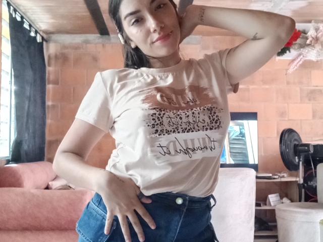 SharonKlart with tan tee, leopard print design, and cursive text, blue jeans, hand on hip, arm raised, smiling, tattoo on arm, brown hair, pink couch in background.
