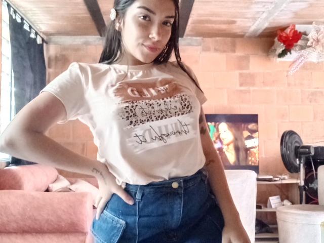 SharonKlart with long brown hair, wearing a white tee showing a print and denim shorts, standing with hand on hip, showing a small tattoo on her right arm.