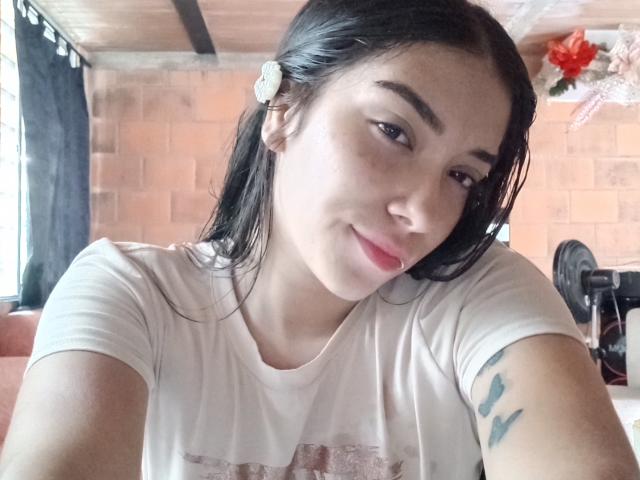 SharonKlart, con i capelli neri bagnati, una camicia bianca, piccoli seni, un tatuaggio sul braccio destro, labbra rosa, un fiore bianco tra i capelli, con un leggero sorriso, che guarda verso la fotocamera, in un ambiente interno..