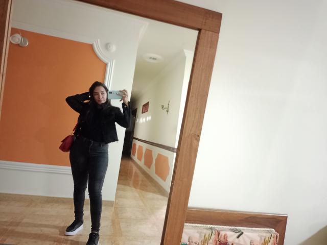 SharonKlart se tient devant un miroir et prend un selfie. Elle porte un haut noir, un jean noir moulant et des baskets noires. Sa main est dans ses cheveux, et elle montre ses fesses..