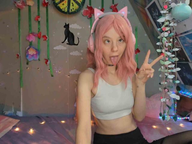 MissAstravert with pink hair, cat ears, and headphones, sticking out her tongue, making a peace sign, wearing a white crop top, showing her small tits, and black skirt.