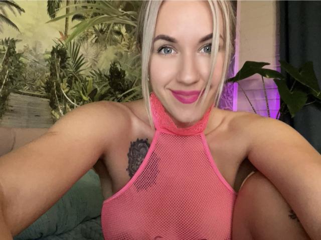 Misstyk with blonde hair, pink mesh halter top, pink lipstick, tattoo on right shoulder, smiling at camera, arms raised, green eyes, pink nipples visible through mesh.