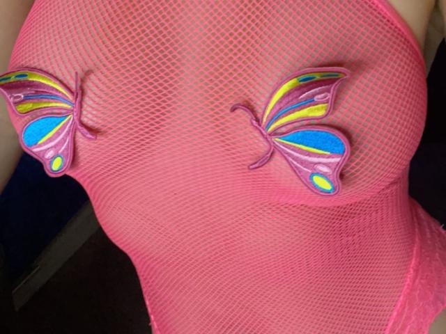 Misstyk wearing a pink fishnet top with colorful butterfly pasties over her tits, showing a bright pink, yellow, blue, and green pattern. Her nipples are covered.