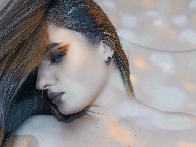 KariMillie's face with long brown hair, orange eye makeup, and dark lipstick, looking down. She's got a small ear piercing and a visible neck. Bokeh light effects.