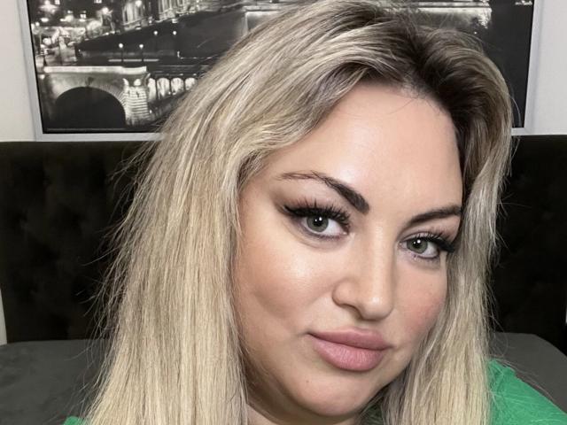 HugeNaturalTits with blonde hair, green eyes, and full pink lips, wearing green, has thick black eyelashes and a slight smirk. Her ass is not visible.