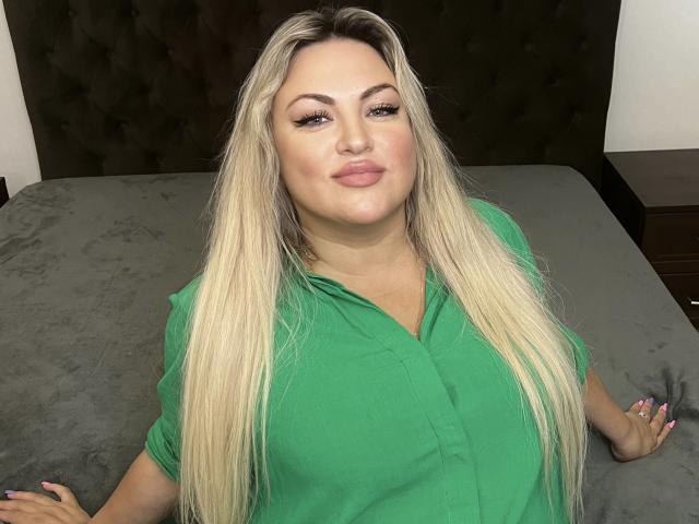HugeNaturalTits with long blonde hair, green shirt, and pink lips, sitting on a gray bed, looking at camera with a slight smile, hands resting on bed.