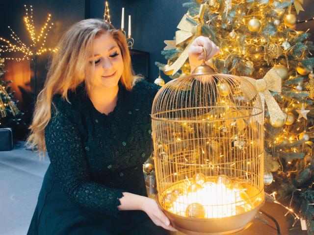 Jessika Daniels, avec ses longs cheveux blonds, portant une robe noire pailletée, tient une cage à oiseaux dorée avec des lumières scintillantes, à côté d'un sapin de Noël décoré..
