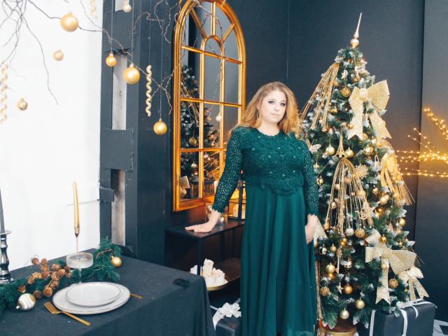 Jessika Daniels steht in einem dunklen Raum, gekleidet in ein grünes, langärmeliges Kleid mit Pailletten, neben einem geschmückten Weihnachtsbaum mit goldenen Ornamenten und Lichtern. Ihr Hintern ist sichtbar..