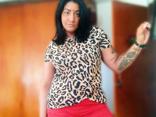 Nahirr with medium brown skin, black hair, wearing a leopard print top, red pants, and tattoos on her right arm, standing in a room, looking at the camera.