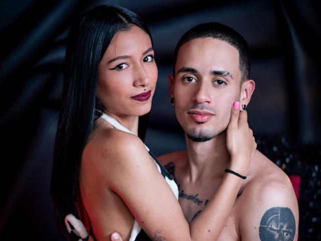 SophiaAndTony, a Latina woman with long black hair and dark lipstick, embraces Tony, a Latino man with tattoos and a shaved head, both shirtless, in a dark room.