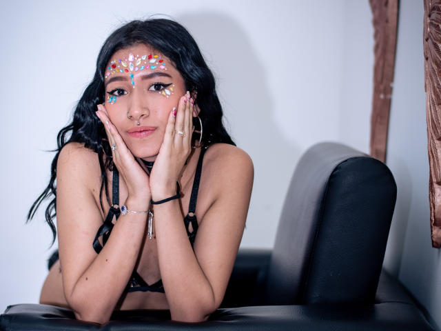 SophiaAndTony with dark hair, colorful face paint, black bra, hands on cheeks, looking at camera, sitting on black leather chair, wearing hoop earrings, bracelet, and rings.