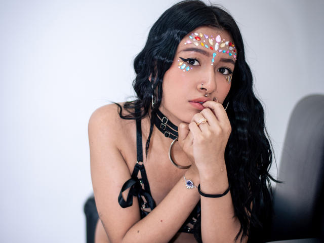 SophiaAndTony with black hair, wearing a black lace bra, choker, and face paint with colorful dots. She's touching her nose, showing a nose ring and ring on her finger.