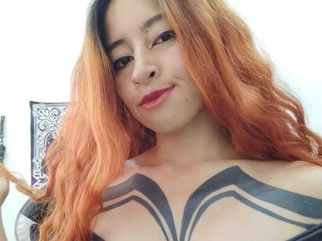 YulikoOfficial con i capelli arancioni, un tatuaggio nero sul petto, che sorride con rossetto rosa, tenendo i capelli sul lato sinistro. Il seno è visibile, con un tatuaggio al centro del petto. Pelle chiara, aspetto casual..