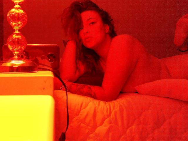 ElectricSapphire, naked, with wavy brown hair, laying on a bed, red-lit, looking at camera, hand on chin, tattoo on arm, white quilt, lamp with glowing orb, plug visible.