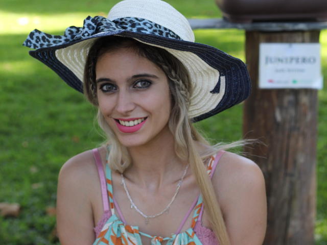 DelaMoons with blonde hair, pink lipstick, and blue eyes, wearing a leopard-print sunhat, colorful dress, and necklace, smiling at the camera, showing her tits.