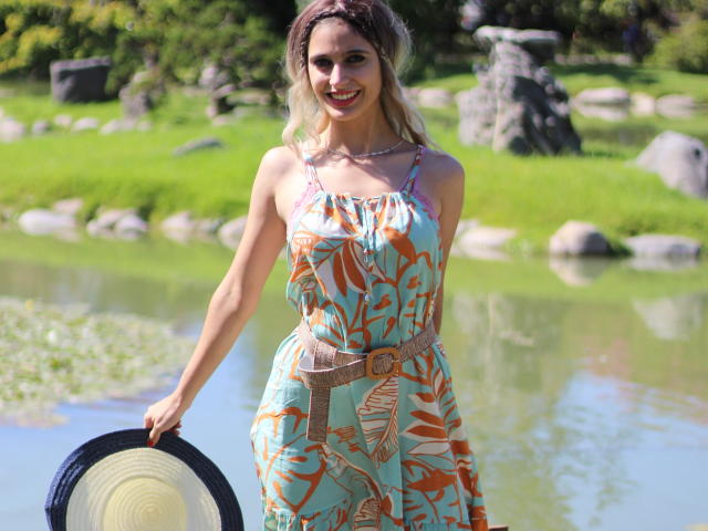 DelaMoons met blond haar, gekleed in een bloemenjurk met oranje en blauwe patronen, die een strohoed vasthoudt, lachend, staand bij een groen grasveld en een reflecterend wateroppervlak..