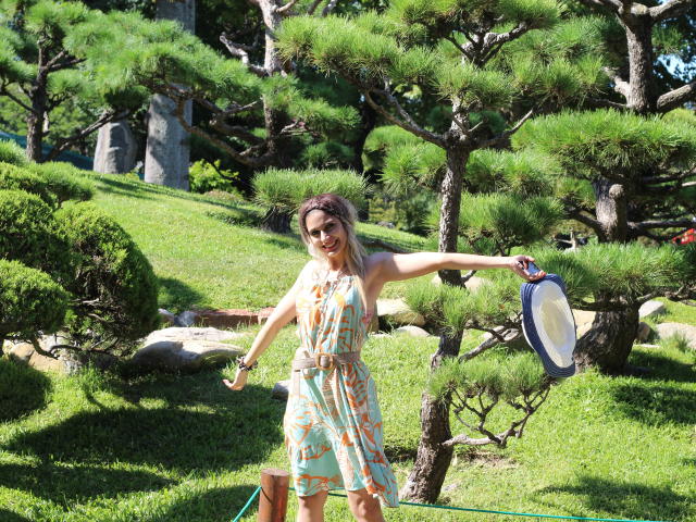 DelaMoons con senos, vestida con un vestido floral sin mangas, sosteniendo un pandereta azul y blanca, con los brazos extendidos, sonriendo, de pie en un jardín soleado y lleno de árboles. Césped verde, roca..
