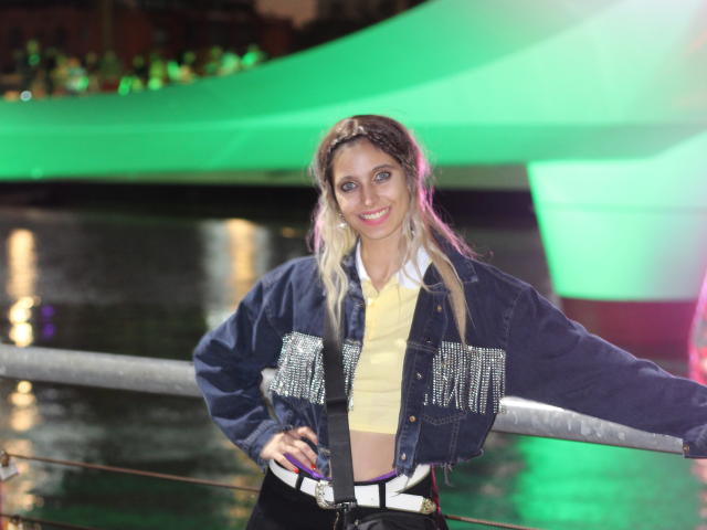 DelaMoons, com cabelos loiros longos, olhos azuis e um sorriso, vestindo uma jaqueta jeans com aplicações brilhantes, uma blusa amarela e calças pretas, apoiada em um corrimão à noite..