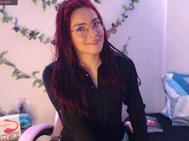 EimmyHoskX has red hair, glasses, and a black shirt, smiling with a cute face, sitting in a pink chair, with a pillow and box in the background.