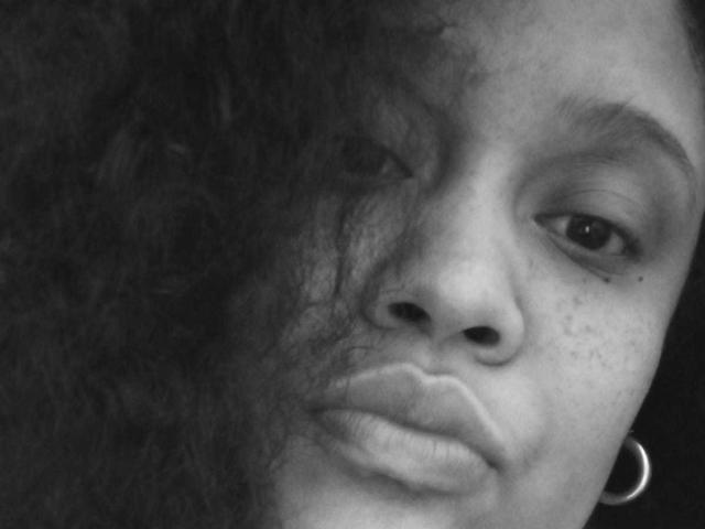 Black-and-white selfie of MillaRoberts69 showing her puffy lips, slightly parted, with curly hair partially covering her right eye, wearing a hoop earring, and freckles on her cheek.