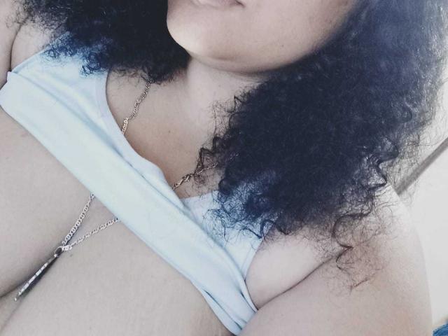 Milla Roberts 69 mit lockigem, schwarzem Haar, heller Haut, die ihre großen Brüste in einem hellblauen Top zeigt, einer silbernen Halskette und einem leichten Lächeln, in einem leicht unscharfen Selfie..