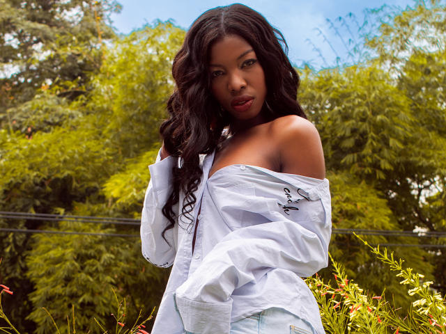 MichelleFlores with long, wavy black hair, wearing a white off-shoulder shirt, exposing her dark, pretty boobs, standing in a grassy area with green trees in the background.