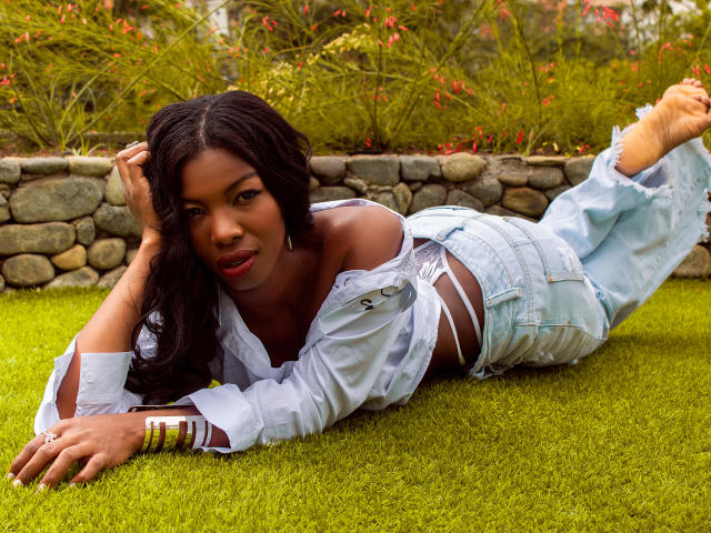 MichelleFlores is lying on grass, her dark tits visible through an open white shirt. She's wearing light blue, ripped jeans and gold bracelets, with long black hair.