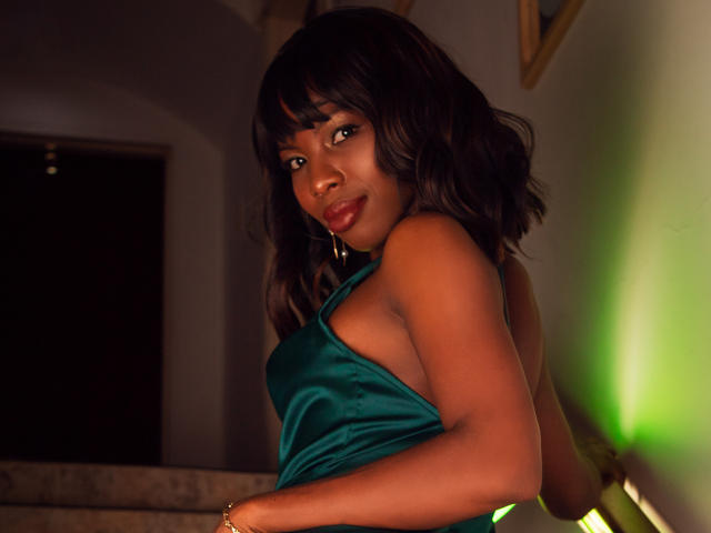 MichelleFlores with dark, wavy hair, wearing a green satin dress, showing her tits, standing in a dimly lit room, green light on the right, smiling at the camera.