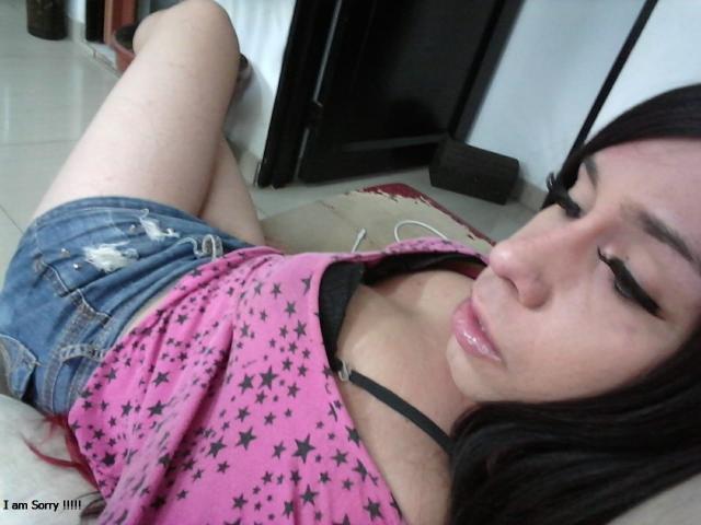 SexyInch lying on a floor, wearing a pink star-patterned top, black bra, and denim shorts, with black eyeliner, and a slightly pouty face, showing her tits.