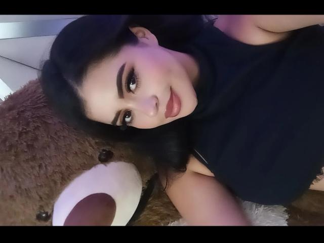 Agatha Vega, con cabello oscuro, cejas pobladas y lápiz labial rosa, está acostada junto a un osito de peluche marrón con una nariz blanca, y lleva una camisa negra. Sus senos son parcialmente visibles..