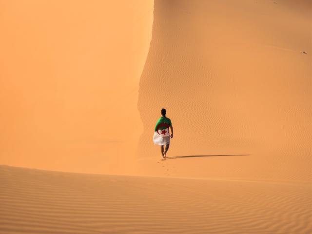 SuperbissimaTroiana, con una camicia verde e pantaloni bianchi, cammina a piedi nudi in un vasto deserto arancione, portando una borsa rossa nella mano destra. Increspature nella sabbia..