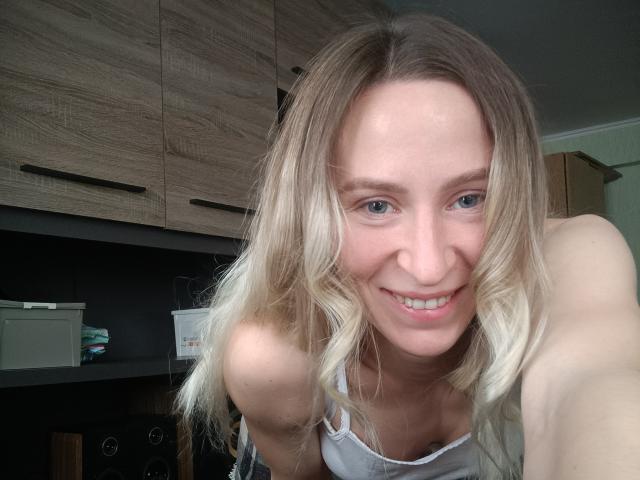 Saveta, con cabello rubio, sonriendo, vestida con una camiseta blanca, inclinándose hacia adelante, mostrando sus senos, y con ojos azules, en una cocina..