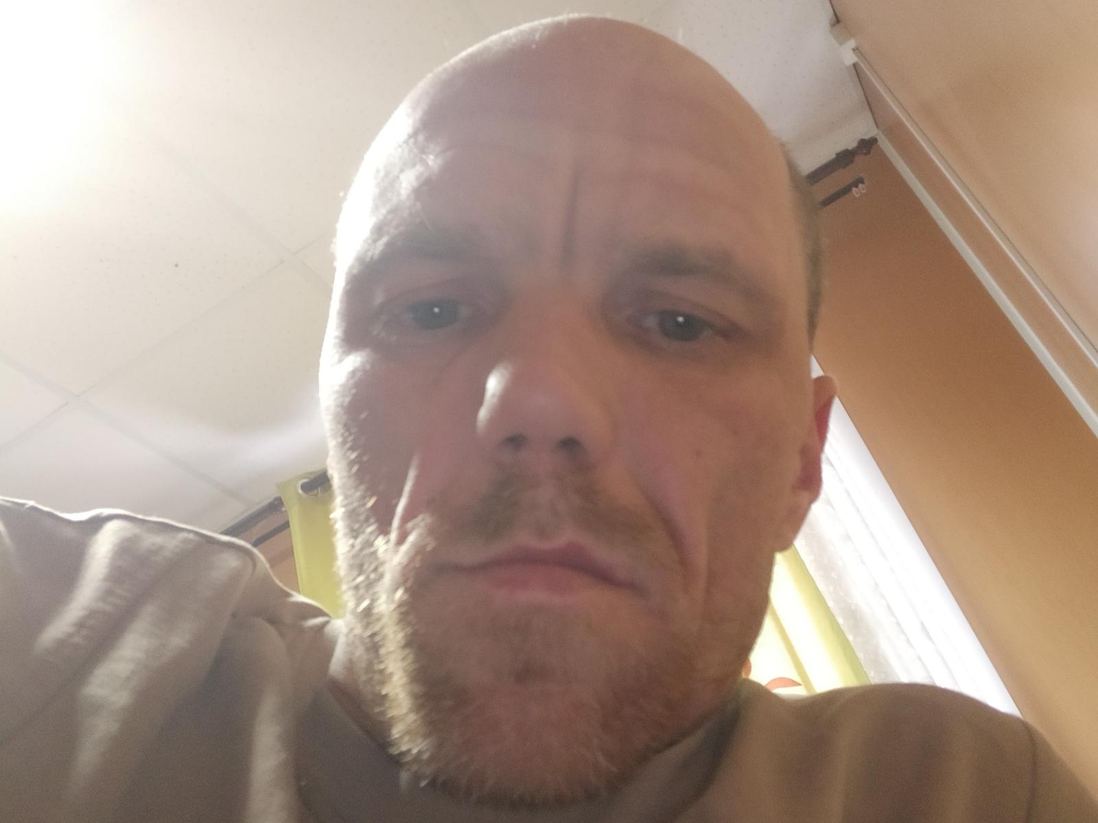 Domovoi careca, com barba, rosto levemente inchado e expressão neutra, vestindo uma camisa marrom, em um selfie tirado de baixo..