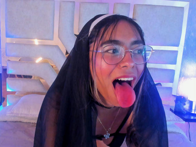 ZafiLeione with black veil, glasses, and tongue out, wearing a white headband, black choker, and necklace, exposing her tits under sheer black fabric. Bedroom with blue lighting.