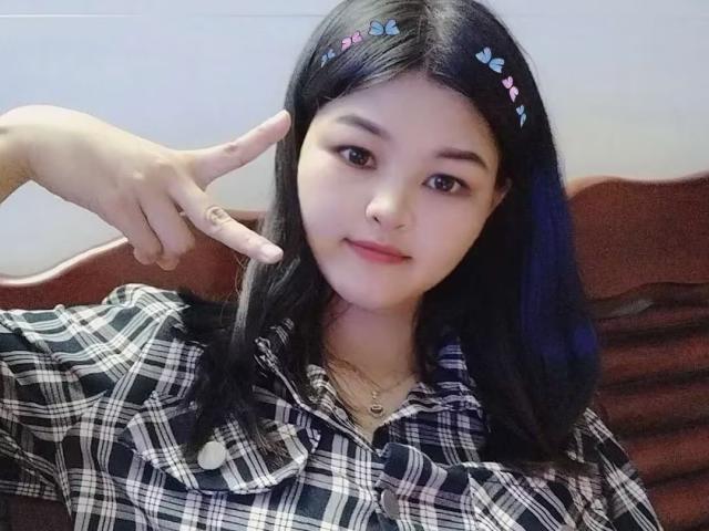 AdaCn with black hair, wearing a plaid shirt, pointing a finger with a peace sign, cute smile, blue butterfly hair clips, and a necklace, sitting on a brown chair.