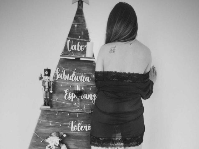 TaniaRey, dos au dos de la caméra, porte un haut noir en dentelle à épaules dénudées et se tient devant un sapin de Noël, montrant un petit tatouage sur son épaule droite..