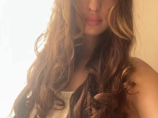 LisaAlisia a de longs cheveux bruns ondulés avec des mèches blondes, elle porte un haut argenté brillant, et ses lèvres pulpeuses sont légèrement visibles dans un autoportrait pris dans une lumière douce et chaude..