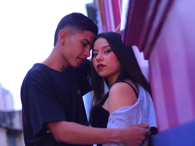 KylieAndJustin with short black hair, black t-shirt, and earring, arms around Justin with long black hair, white off-shoulder top, black bra, and red lipstick, in an urban setting.
