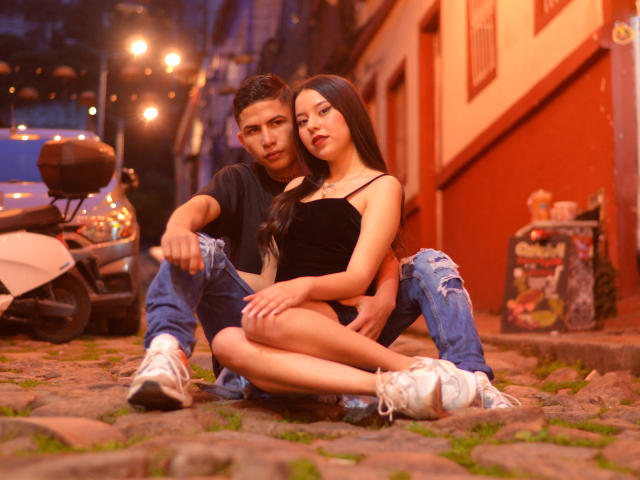 KylieAndJustin, with long black hair and a black dress, sits on a cobblestone street with Justin, wearing ripped jeans and white sneakers, his arm around her.