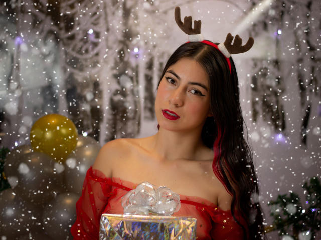 JuliaGarciax with red sheer top, brown antler headband, holding gift with clear ribbon, red lipstick, dark hair, snow effect, golden balloon in background. Boobs partially visible.