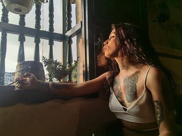 EmaPloxy, con lunghi capelli ricci castani, indossa un top corto bianco che mette in mostra un seno abbondante e tatuaggi, è appoggiata al vetro di una finestra, fumando, con la luce del sole sul viso, una finestra di legno e una pianta in vaso all'esterno..