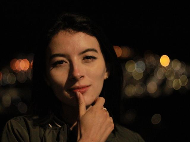 GabrielaMoore with dark hair, slightly smiling, finger touching her chin, dimly lit, bokeh lights in background, wearing a green shirt, subtle makeup, ring on her finger.