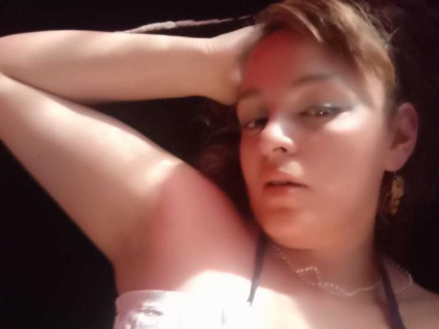 AmandaHotL with short brown hair, smoky eye makeup, white top, arm raised, looking at camera, red light on cheek, necklace, and gold earring, in dark setting.