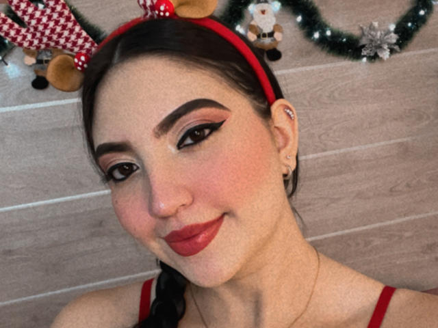 Megan Hils, avec du rouge à lèvres, des sourcils noirs épais et un serre-tête rouge avec des oreilles de renne, portant un haut rouge, a un joli sourire et montre son nez..