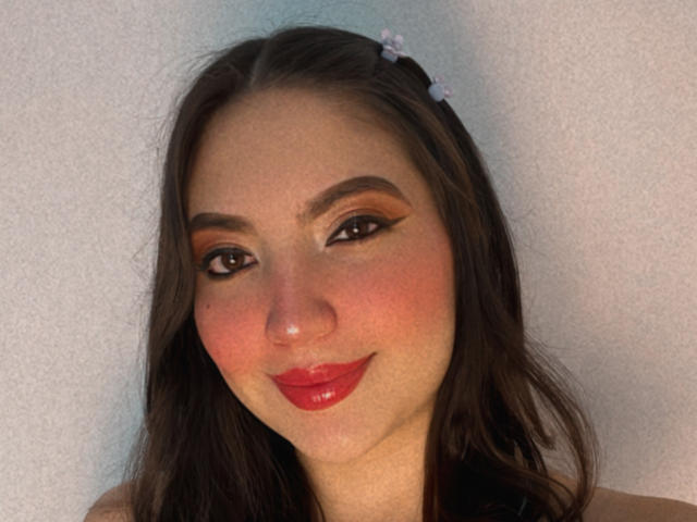 MeganHils tem batom vermelho, delineador escuro e olhos castanhos com um sorriso discreto. Ela tem cabelos castanhos longos com pequenas flores brancas e um rosto levemente salpicado de sardas..